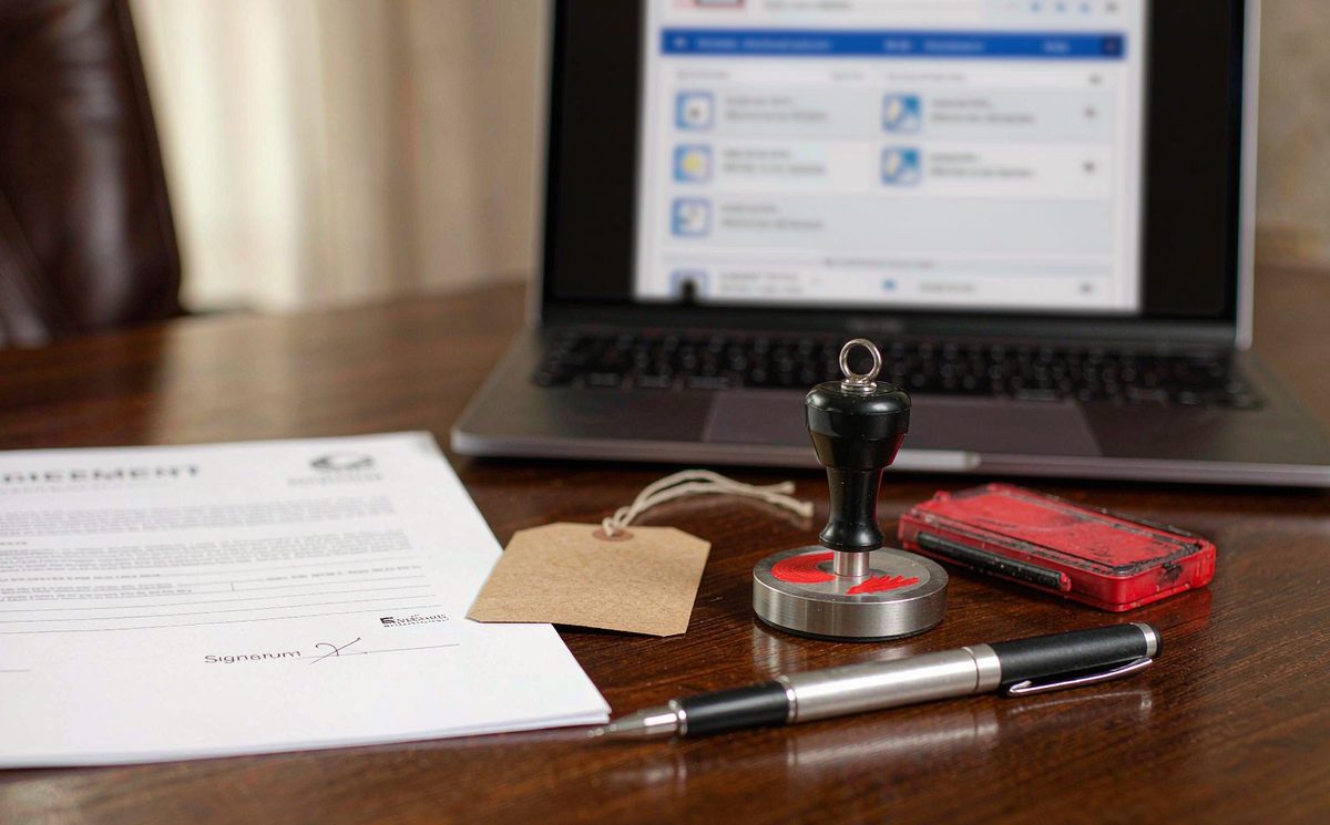 A contract on a desk with a fountain pen, an ink stamp and a blank clothing hang-tag, a trademark search on a laptop behind